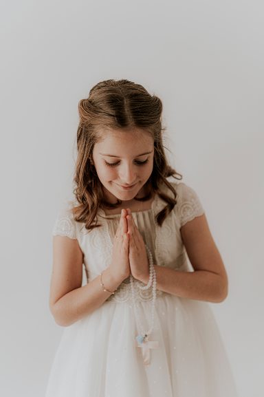 Niña con cabello castaño claro, vestida de blanco y con las manos juntas en actitud de oración.