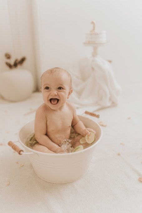 Bebé sonriente sentado en una tina blanca con agua, rodeado de decoraciones suaves.