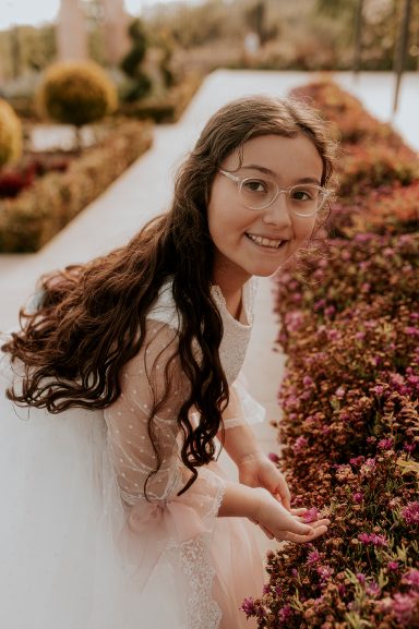 Niña sonriendo entre flores en un jardín, con cabello largo y ondulado.
