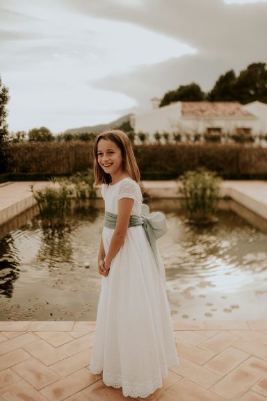Niña con vestido blanco y cinta verde, de pie junto a una fuente en un paisaje natural.