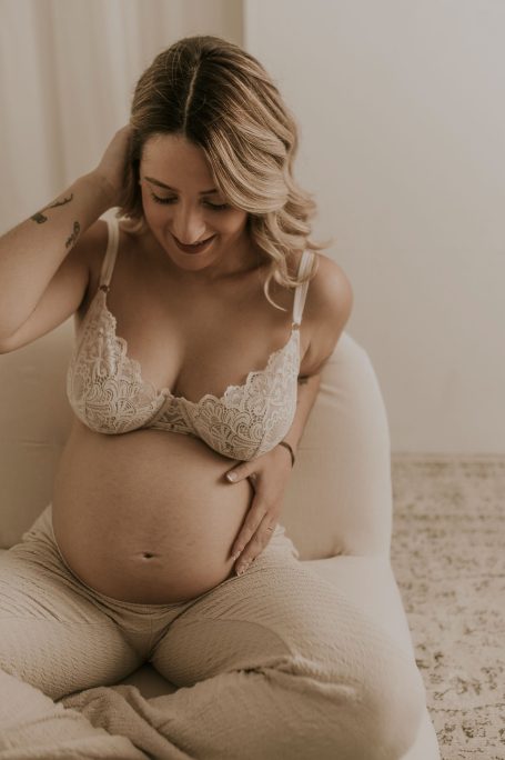 Mujer embarazada sonriendo, acariciándose la barriga, vestida con lencería y pantalones claros.