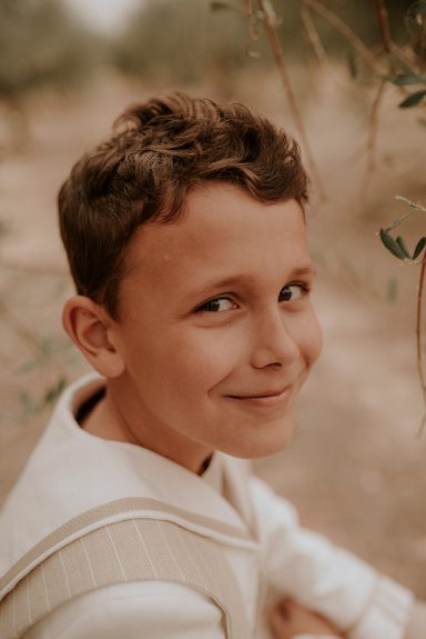 Niño con cabello castaño claro sonriendo, rodeado de naturaleza.