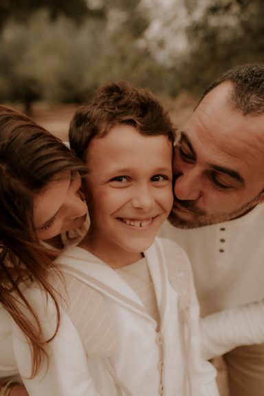 Niño sonriendo entre dos adultos, recibiendo cariño en un entorno natural.