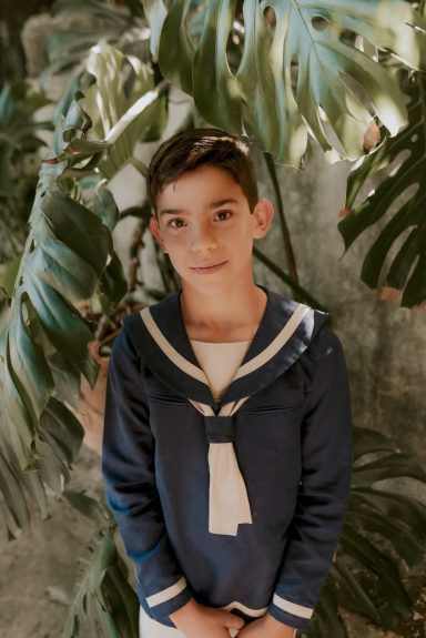 Niño con un uniforme marinero azul frente a plantas verdes.