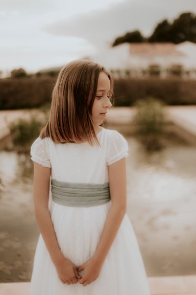 Niña de espaldas, con vestido blanco y cinta verde, frente a un estanque.