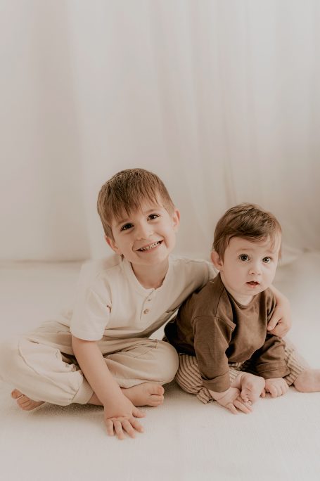 Niños sentados en el suelo, uno sonríe y el otro los observa, con ropa de tonos suaves.