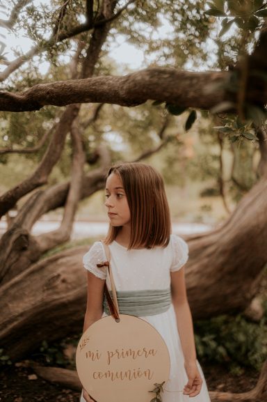 Niña con un vestido blanco y cinta verde, sostiene un cartel en un entorno natural.