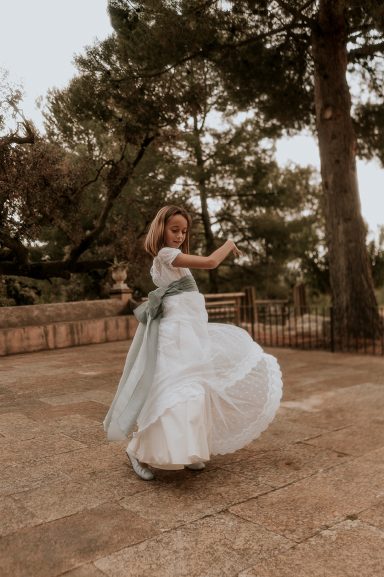Niña girando con un vestido blanco y cinta celeste, rodeada de árboles.