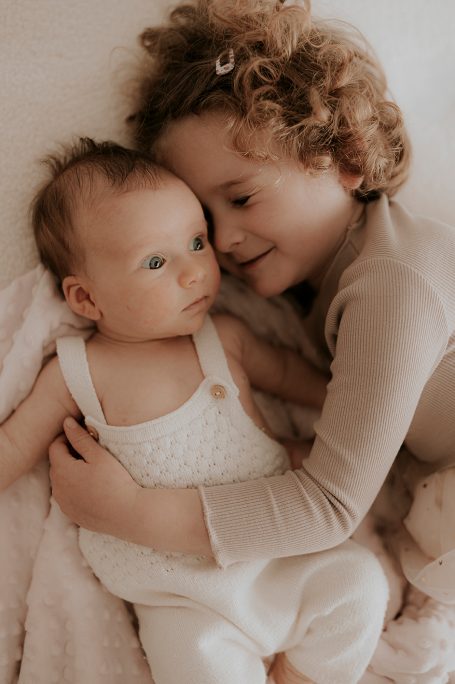 Niña abrazando a un bebé sobre una manta clara, ambos lucen felices y tranquilos.