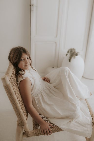 Niña sentada en una silla, vistiendo un vestido blanco con fondo claro y decoraciones suaves.