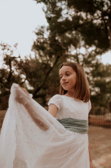 Niña con vestido blanco y cinta azul, sonriendo mientras agita una tela.