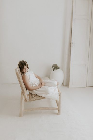 Persona sentada en una silla de madera, vestida de blanco, en un entorno minimalista.