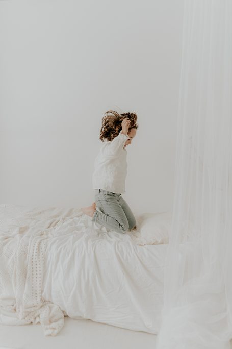 Niña saltando en una cama blanca, con un ambiente suave y luminoso.
