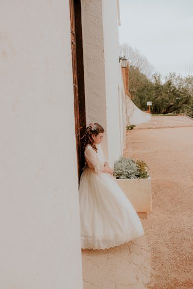 Niña con vestido de novia, de pie junto a una pared exterior. Fondo natural.