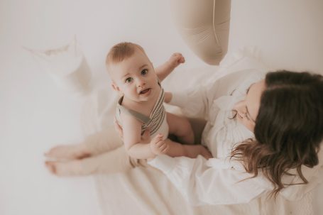 Bebé sonriente sentado en una sábana blanca junto a una madre cariñosa.