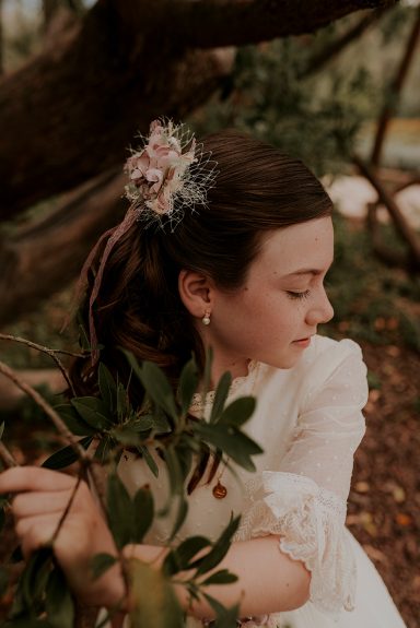 Niña con vestido blanco y peinado adornado con flores, rodeada de ramas y hojas.