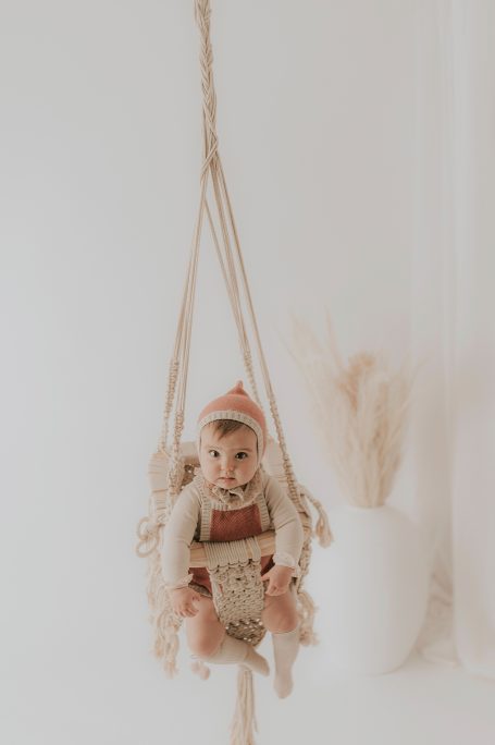 Un bebé en un columpio con un libro en sus manos, vestido con un gorro de punto.