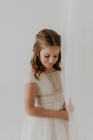 Niña con vestido claro, mirando hacia abajo, junto a una cortina blanca.