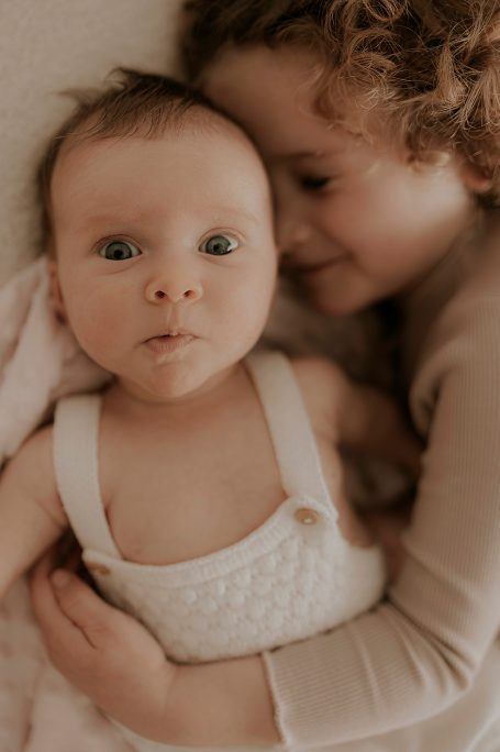 Bebé en un mono blanco con una niña abrazándola, ambos sonriendo.