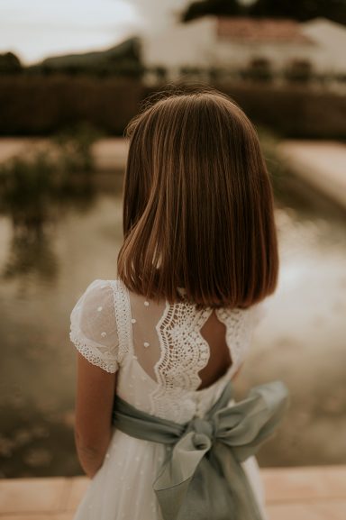 Niña de espaldas con vestido blanco y lazo azul, frente a un estanque.