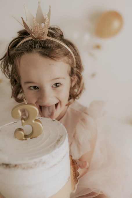 Niña sonriente con una corona, sosteniendo un pastel decorado con el número 3.