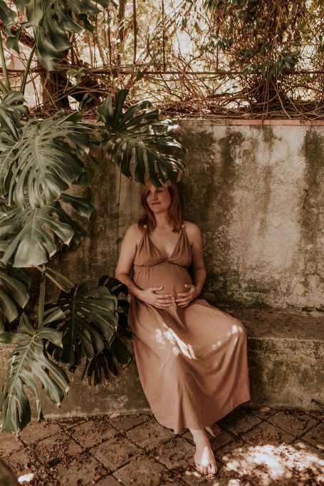 Mujer embarazada sentada junto a plantas verdes en un entorno natural y cálido.
