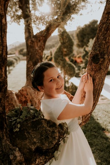 Niña con vestido blanco posando junto a un árbol en un paisaje natural.