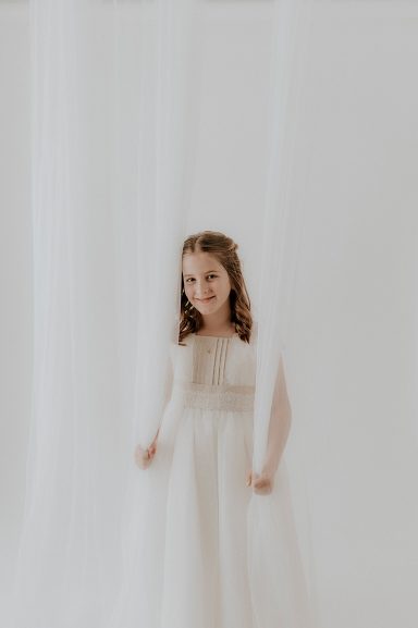 Niña con vestido blanco de pie detrás de una cortina blanca.