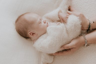 Bebé dormido vestido con un abrigo blanco, sostenido por manos cuidadosas.